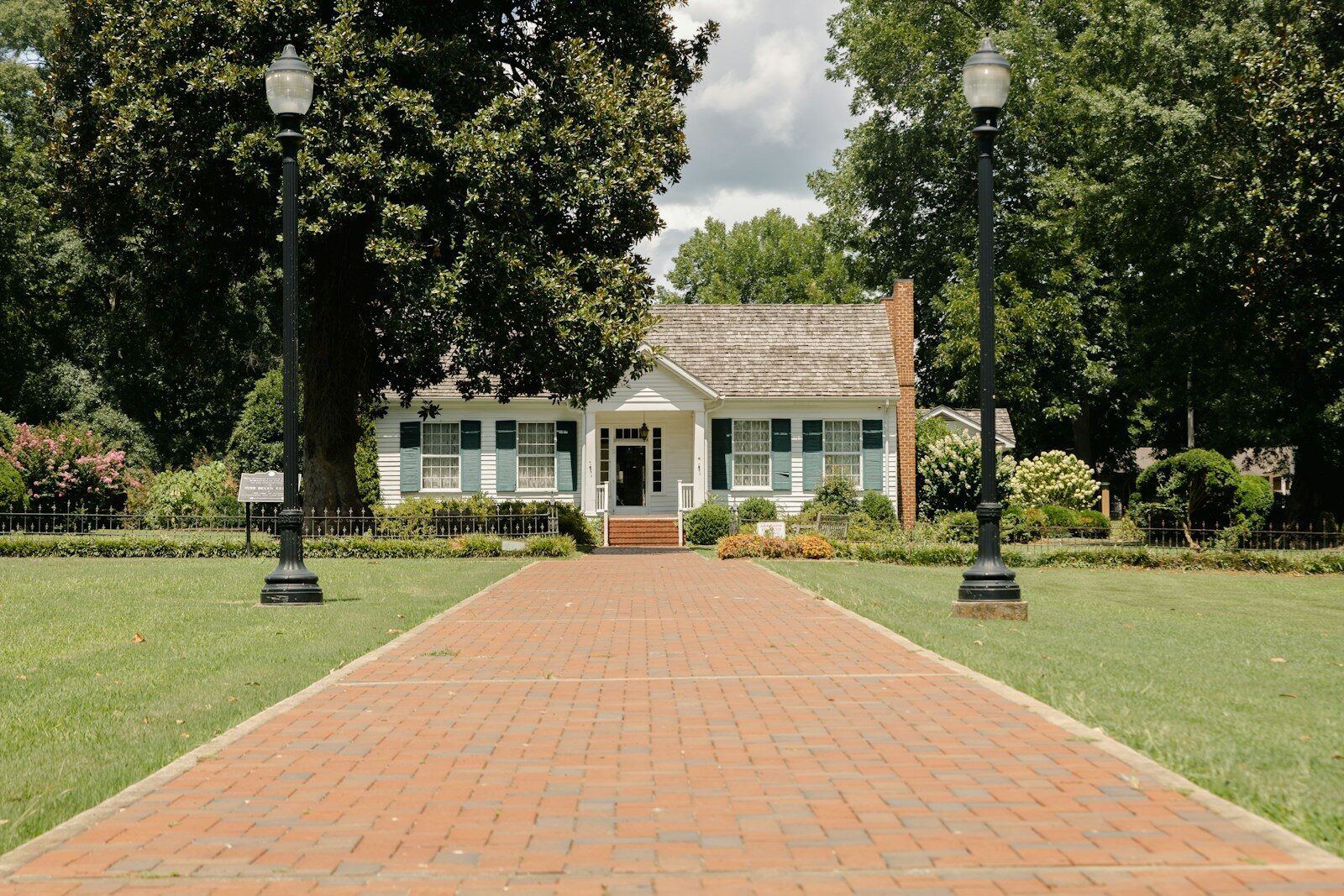 a brick walkway leading to a white house with trees and a lamp post